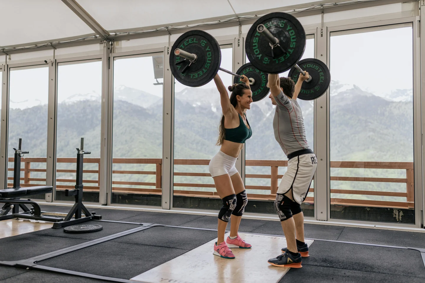 Two lifters pressing barbells overhead in a gym with mountain views